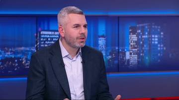 Polityk Adrian Zandberg siedzi w studiu telewizyjnym, m&oacute;wiąc do kamery. W tle widoczne są ekrany z nocnym widokiem na budynki miejskie.