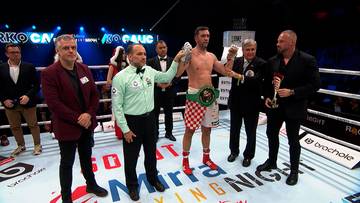 Pięściarz w pasie mistrzowskim stoi na ringu otoczony przez sędziów i promotorów.