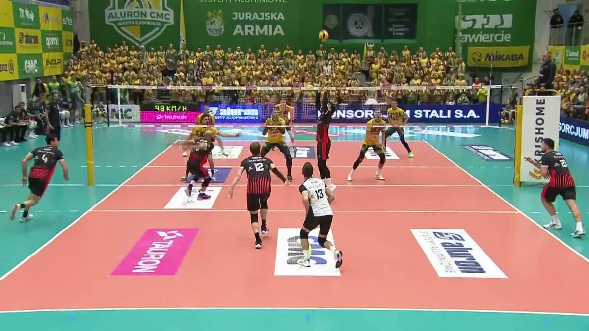 Siatkarze w akcji podczas meczu, jeden z zawodnik&oacute;w wykonuje atak nad siatką, inni zawodnicy pr&oacute;bują blokować.
