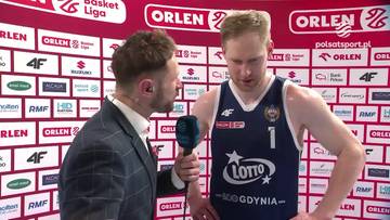Jarosław Zyskowski, koszykarz, udziela wywiadu dziennikarzowi z mikrofonem. Zawodnik ubrany jest w niebieski str&oacute;j sportowy z numerem 7 i logo LOTTO Gdynia. Tło stanowi ściana z białych cegieł z czerwonymi napisami i logo sponsora ORLEN.