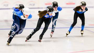 Cztery osoby na łyżwach szybkich ścigają się na torze, wszystkie w kaskach i sportowych strojach.