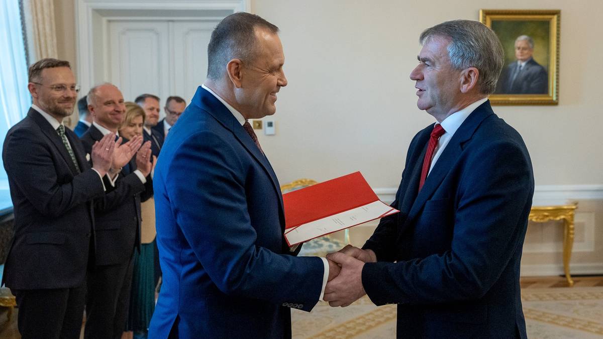 Dw&oacute;ch mężczyzn w garniturach stoi naprzeciwko siebie, jeden wręcza drugiemu dokumenty.