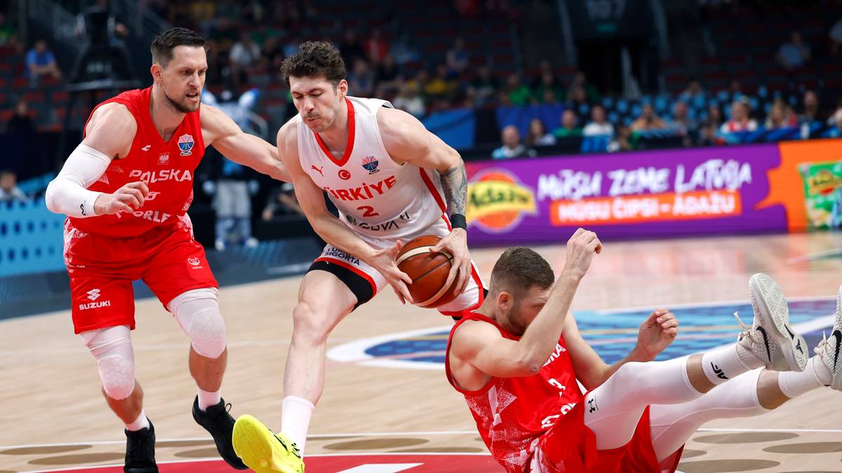 Trzech zawodników drużyny koszykarskiej walczy o piłkę na boisku podczas meczu. Dwóch zawodników w czerwonych strojach próbuje odebrać piłkę zawodnikowi w białym stroju z numerem 2.