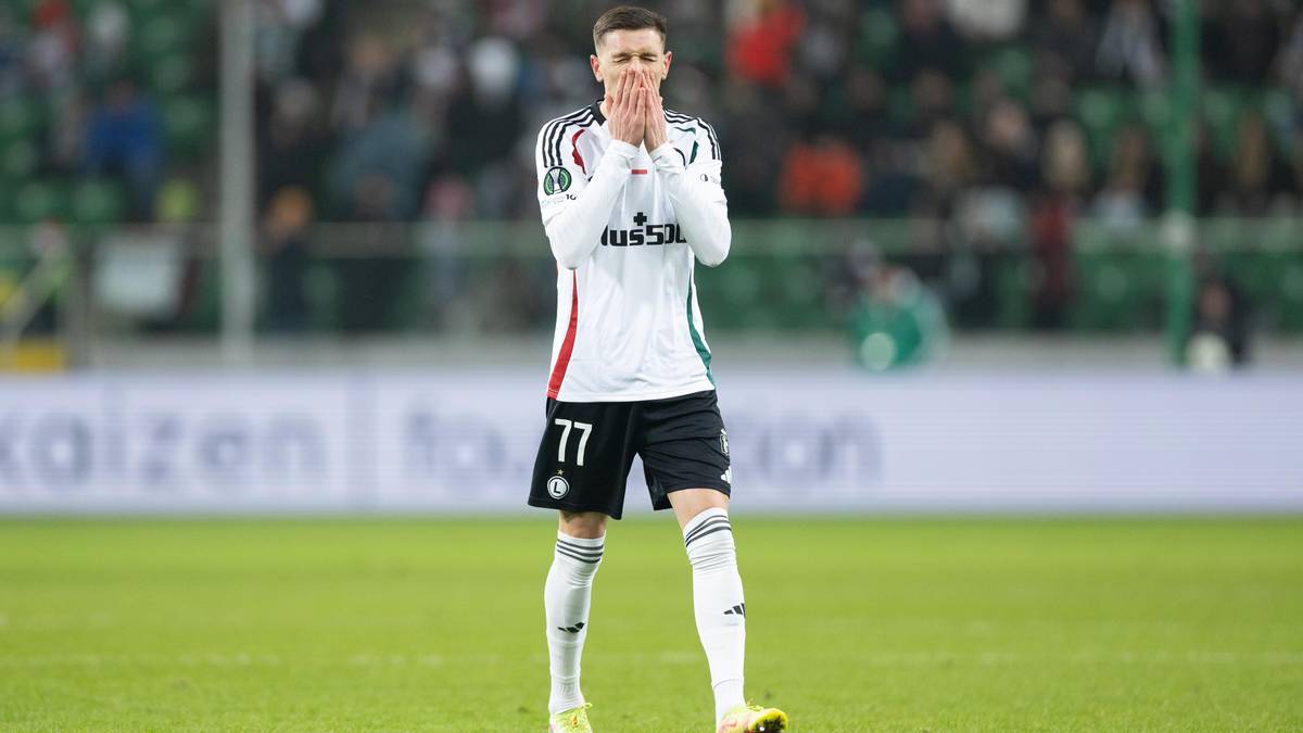 Piłkarz Legii Warszawa w białej koszulce z numerem 77 i czarnych spodenkach stoi na boisku piłkarskim, trzymając dłonie przy twarzy, wyrażając emocje.