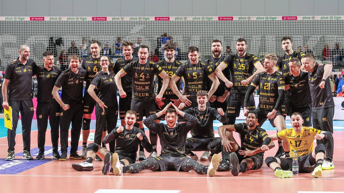 Grupa mężczyzn w strojach siatkarskich czarno-żółtych pozuje do zdjęcia na boisku do siatkówki.