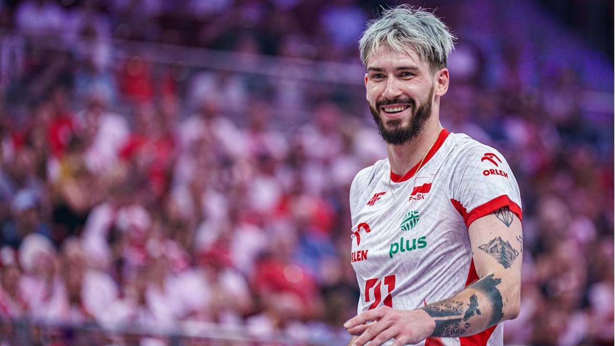 Siatkarz w białej koszulce z czerwonymi elementami, z widocznymi tatuażami, uśmiecha się do widza. W tle rozmyta publiczność.