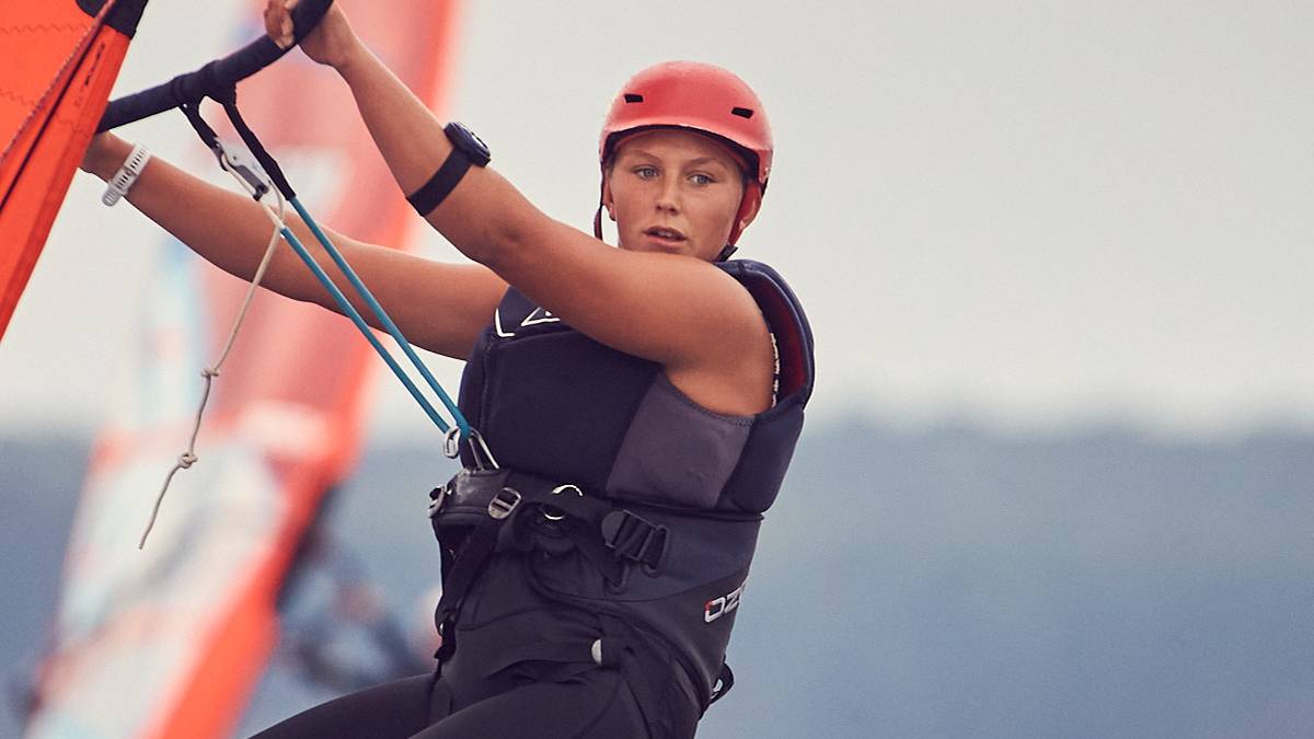 Młoda kobieta w czerwonym kasku i czarnym stroju żeglarskim na desce windsurfingowej, zmagająca się z wiatrem.
