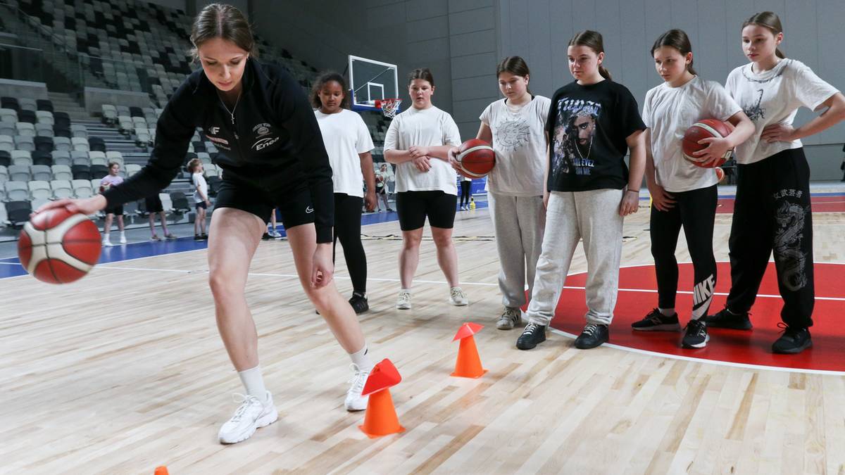 Grupa dziewcząt ćwiczy z piłką do koszykówki na sali gimnastycznej, prowadzone przez jedną z nich.