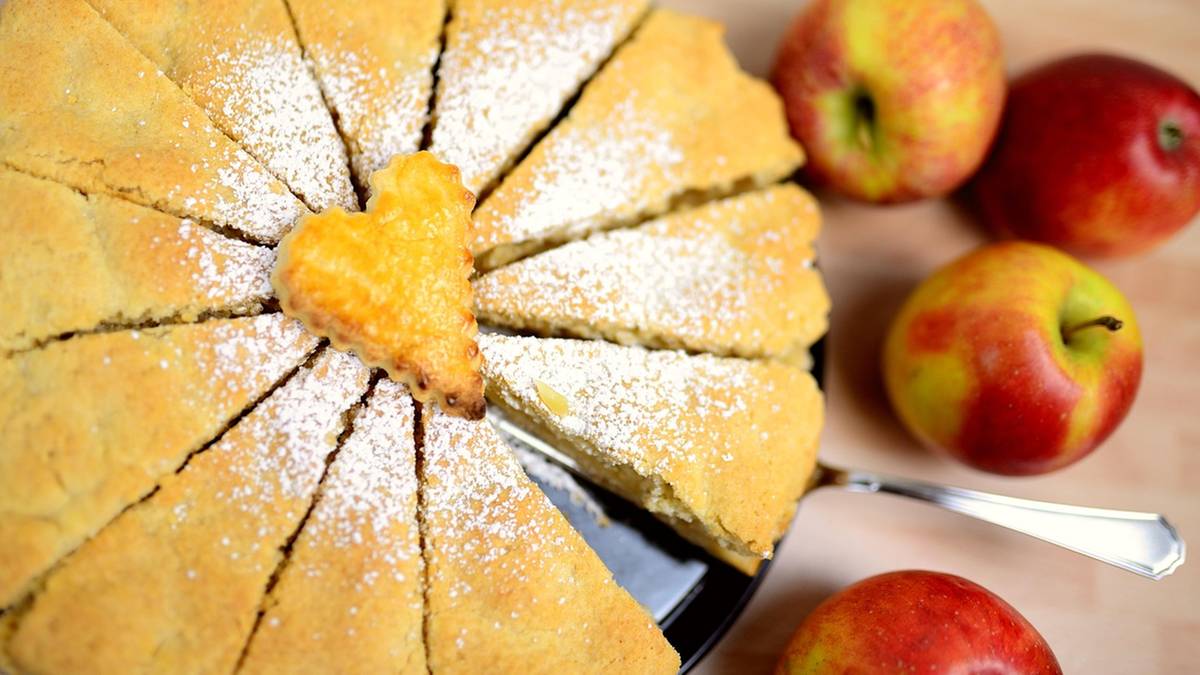 Pokrojone ciasto na kształt koła posypane cukrem pudrem, z wyciętym sercem z ciasta na środku, obok znajdują się jabłka.