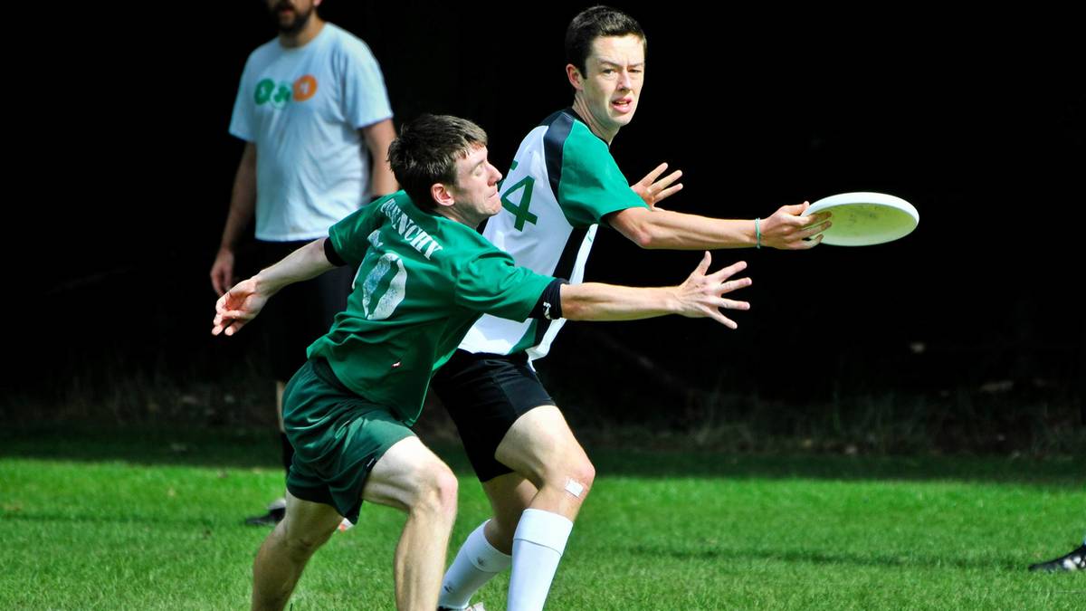Dwóch młodych mężczyzn grających w ultimate frisbee. Jeden z nich, ubrany na zielono, próbuje złapać lecący dysk. Drugi zawodnik, w zielono-białej koszulce, jest w trakcie rzutu dyskiem.