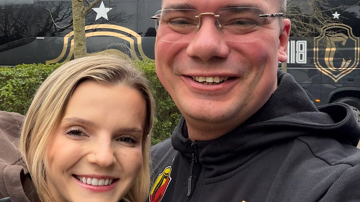 Kobieta i mężczyzna pozują do zdjęcia selfie, oboje uśmiechnięci. Mężczyzna ma na sobie czarną bluzę z kapturem, a za nimi widoczny jest autokar z logo drużyny.