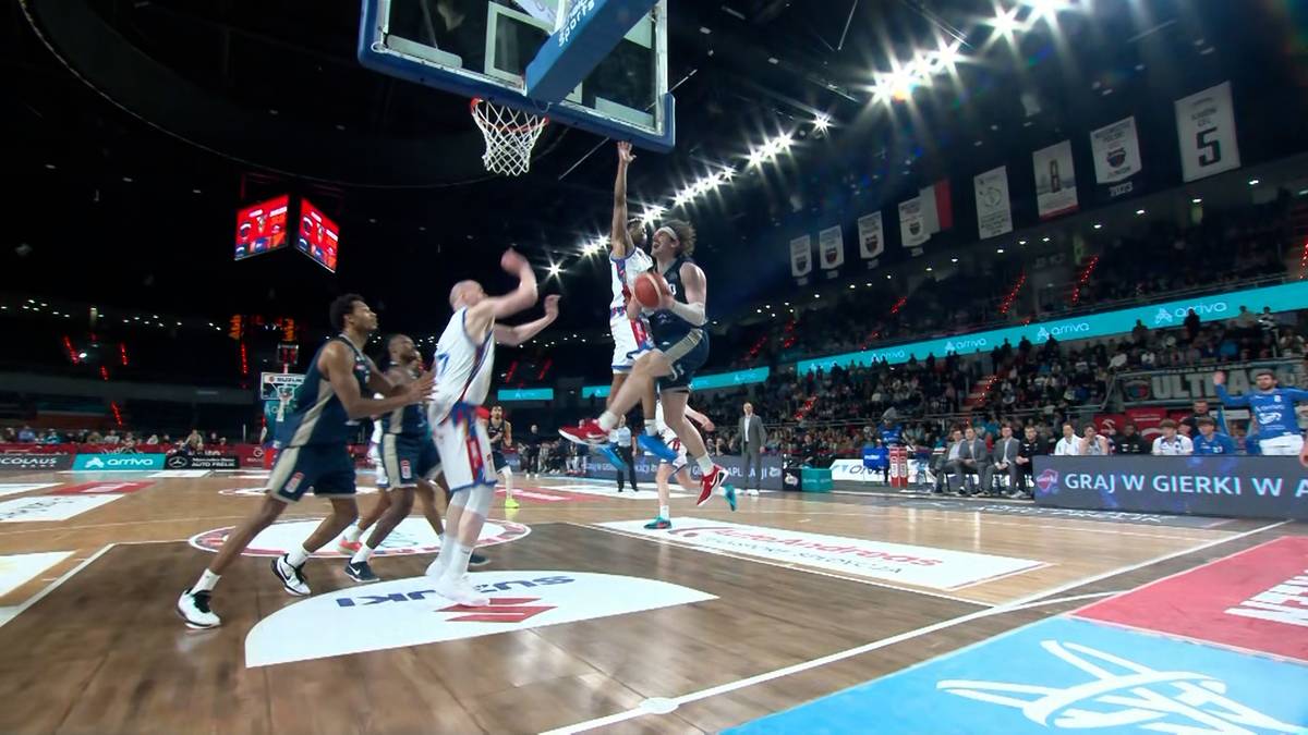 Gracz w niebieskim stroju wykonuje rzut do kosza, podczas gdy dwaj gracze w białych strojach pr&oacute;bują go zatrzymać, na tle publiczności i parkietu do koszyk&oacute;wki.
