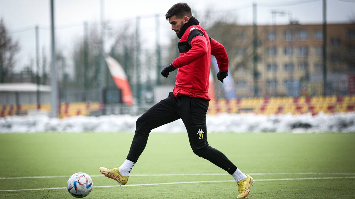 Piłkarz w czerwonej bluzie i czarnych spodenkach wykonuje drybling z piłką na boisku piłkarskim.