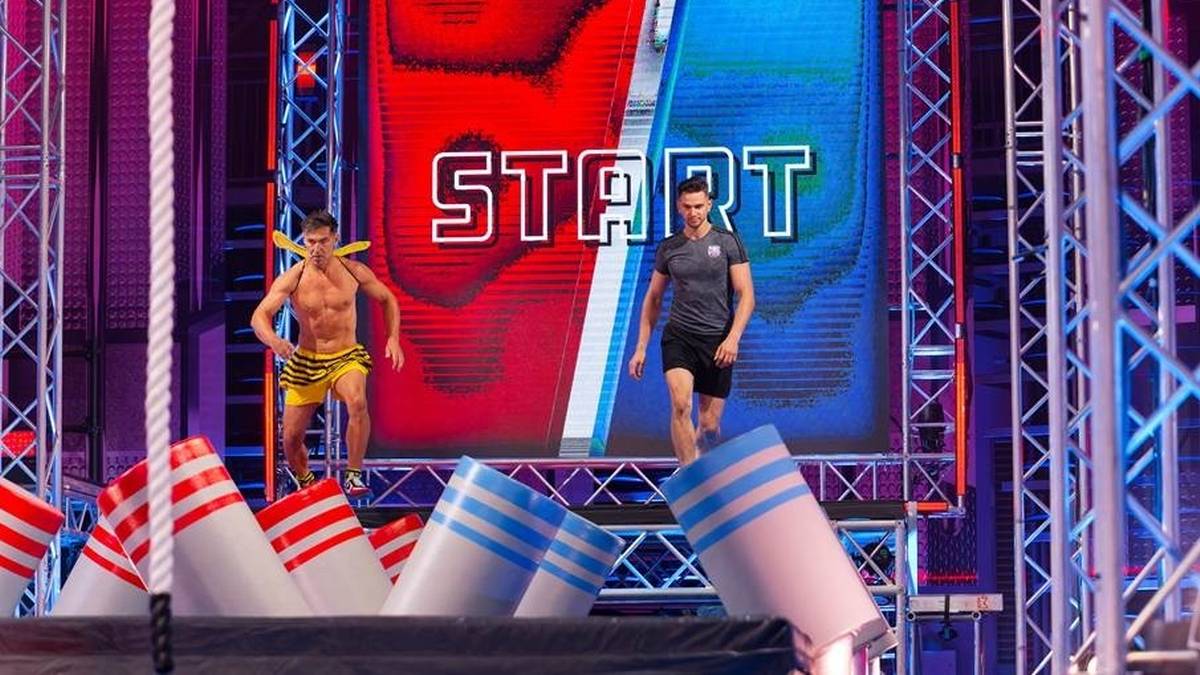 Dwóch mężczyzn gotowych do rozpoczęcia biegu na torze przeszkód "Ninja vs Ninja" przed dużym ekranem z napisem "START".