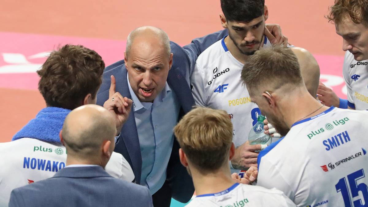 Trener siatkówki w niebieskiej koszuli i marynarce gestykuluje do swoich zawodników, którzy skupieni go słuchają.