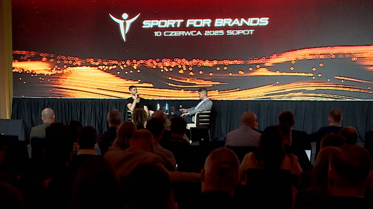 Na scenie podczas konferencji Sport for Brands 2025 siedzą dwie osoby przy stole, a za nimi na dużym ekranie wyświetla się logo wydarzenia z datą i miejscem.