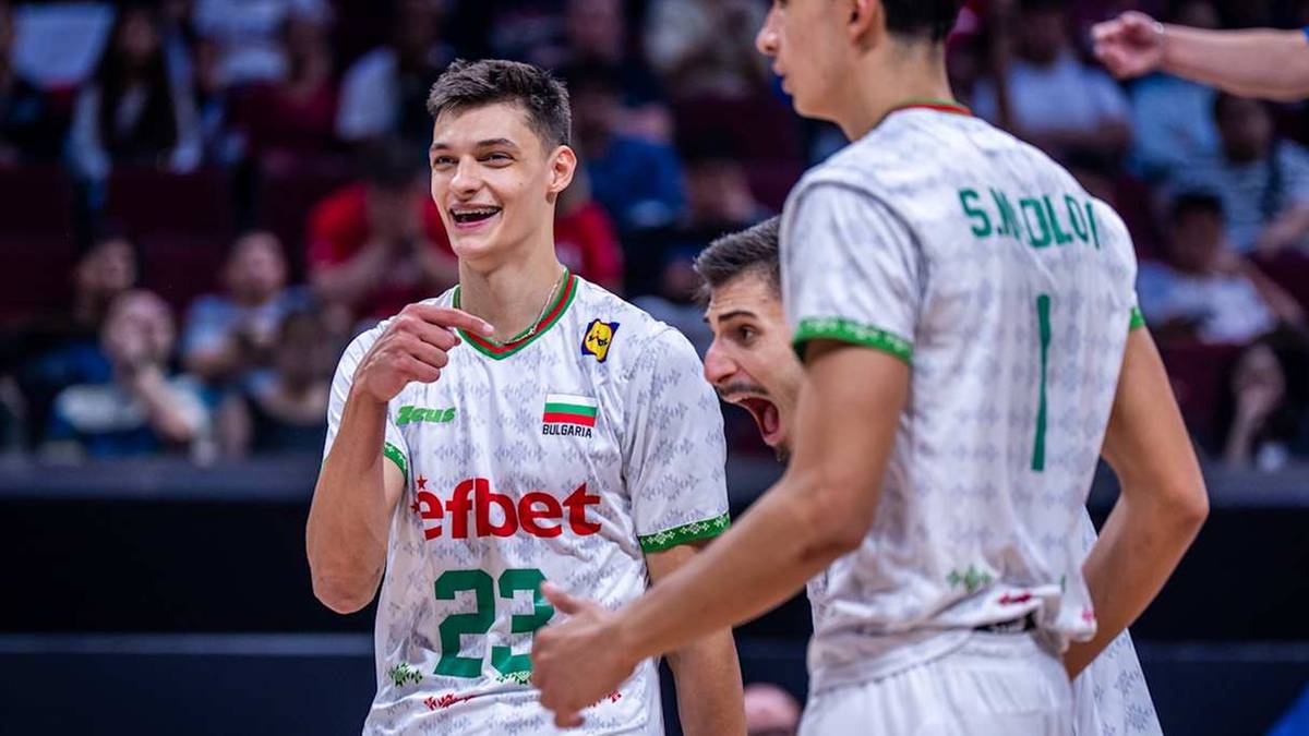 Siatkarz w białej koszulce z numerem 22 i zielonymi wstawkami wskazuje palcem w kierunku widza, uśmiechając się. Obok niego stoją inni zawodnicy w podobnych strojach.