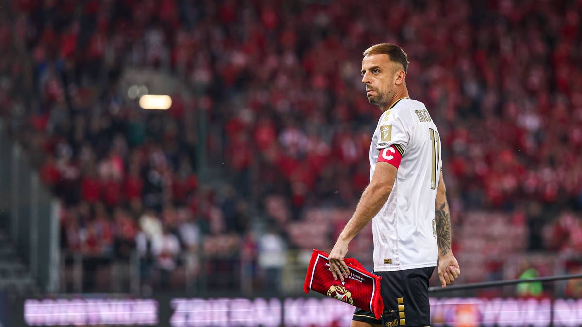 Piłkarz reprezentacji Polski w białej koszulce, stojący na tle trybun wypełnionych kibicami w czerwonych barwach.