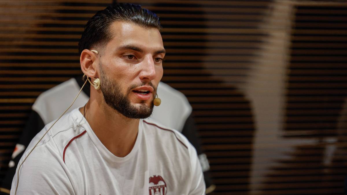 Portret mężczyzny o ciemnych włosach i zarostu, ubranego w białą koszulkę z emblematem. Widoczne są przetworzone słuchawki na jego uszach, sugerujące wywiad lub nagranie.