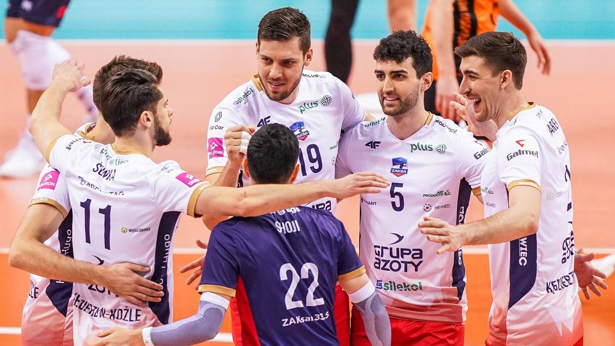 Grupa siatkarzy w białych koszulkach z numerami na plecach świętuje zwycięstwo, jeden z zawodnik&oacute;w w fioletowej koszulce stoi tyłem.