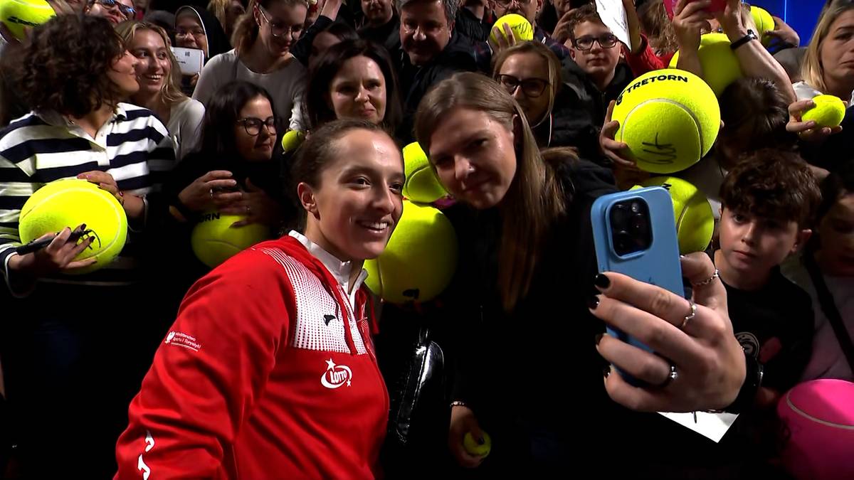 Iga Świątek pozuje do zdjęcia z fanką, która trzyma telefon komórkowy. W tle widać tłum kibiców, z których niektórzy mają piłki tenisowe.