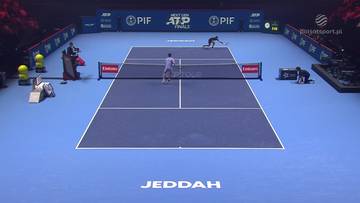 Dw&oacute;ch zawodnik&oacute;w grających w tenisa na niebieskim korcie, jeden z nich po lewej stronie siatki, drugi po prawej. W tle widać bandy reklamowe i trybuny.