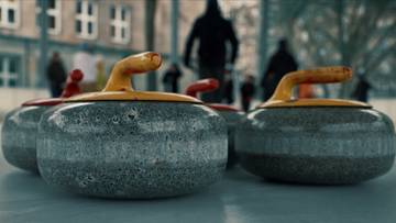 Cztery kamienie do curlingu z ż&oacute;łtymi uchwytami na lodowisku. W tle rozmyte sylwetki ludzi.