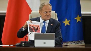Donald Tusk siedzi przy biurku i prezentuje kartkę z rysunkiem na tle flag Polski i Unii Europejskiej.