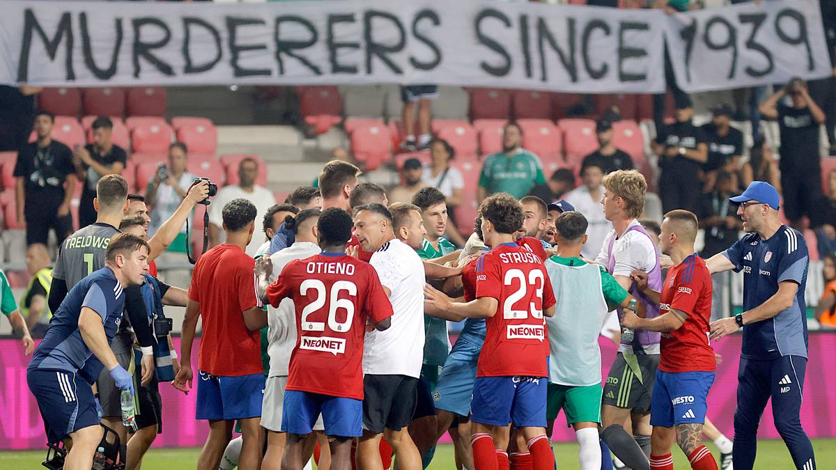 Zgromadzenie kibiców na stadionie piłkarskim, na którym wywieszono transparent z napisem "MURDERERS SINCE 1939". Grupka mężczyzn w strojach sportowych otacza innych kibiców, część z nich wznosi ręce.