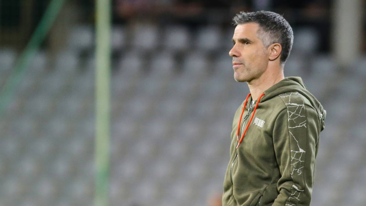 Mężczyzna w ciemnozielonej bluzie z kapturem stoi na tle trybun stadionu.