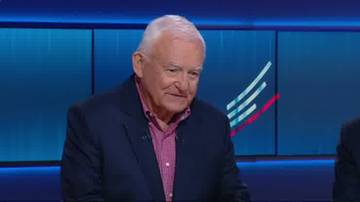 Starszy mężczyzna w ciemnej marynarce i r&oacute;żowej koszuli patrzy w bok z lekkim uśmiechem. W tle widać niebieskie panele z białymi i czerwonymi liniami tworzącymi wz&oacute;r.