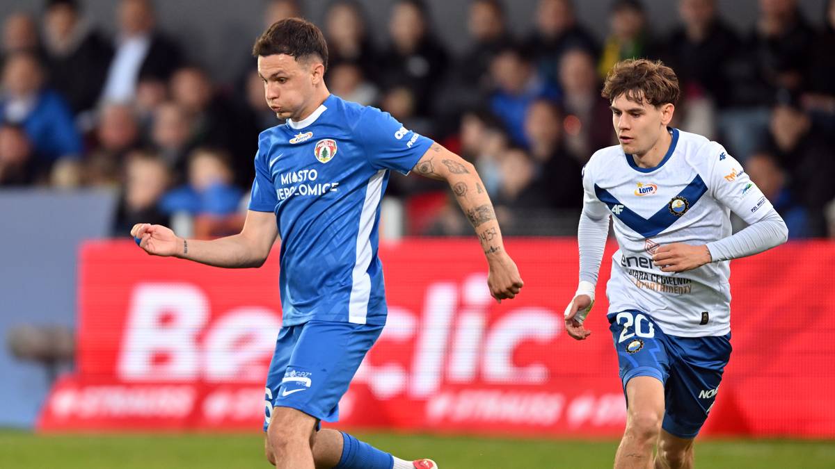 Dwóch piłkarzy w niebieskich i białych strojach rywalizuje o piłkę na boisku piłkarskim, z publicznością w tle.