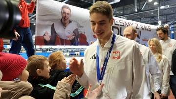 Młody panczenista Semirunnij w białej kurtce z polską flagą, uśmiecha się i wita z kibicami, wśr&oacute;d kt&oacute;rych są dzieci. W tle widoczny jest baner z jego zdjęciem.