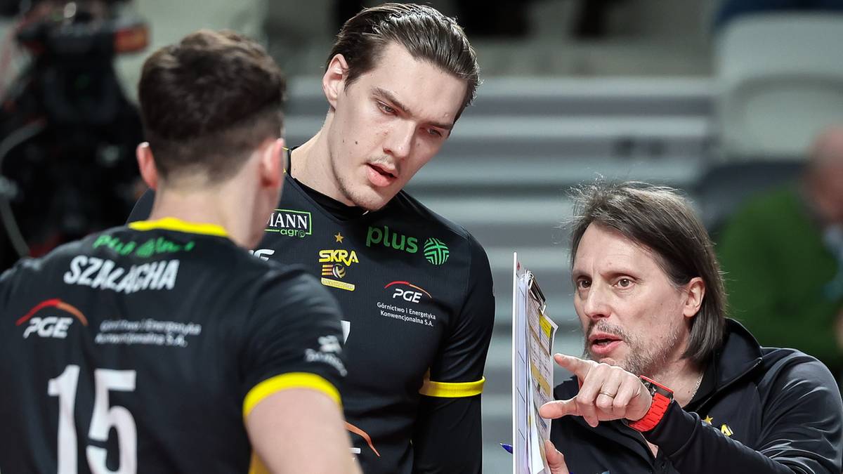 Trzech mężczyzn w strojach siatkarskich podczas rozmowy taktycznej. Dw&oacute;ch młodszych stoi tyłem do widza, jeden starszy, ubrany w ciemną bluzę, pokazuje coś na kartce.