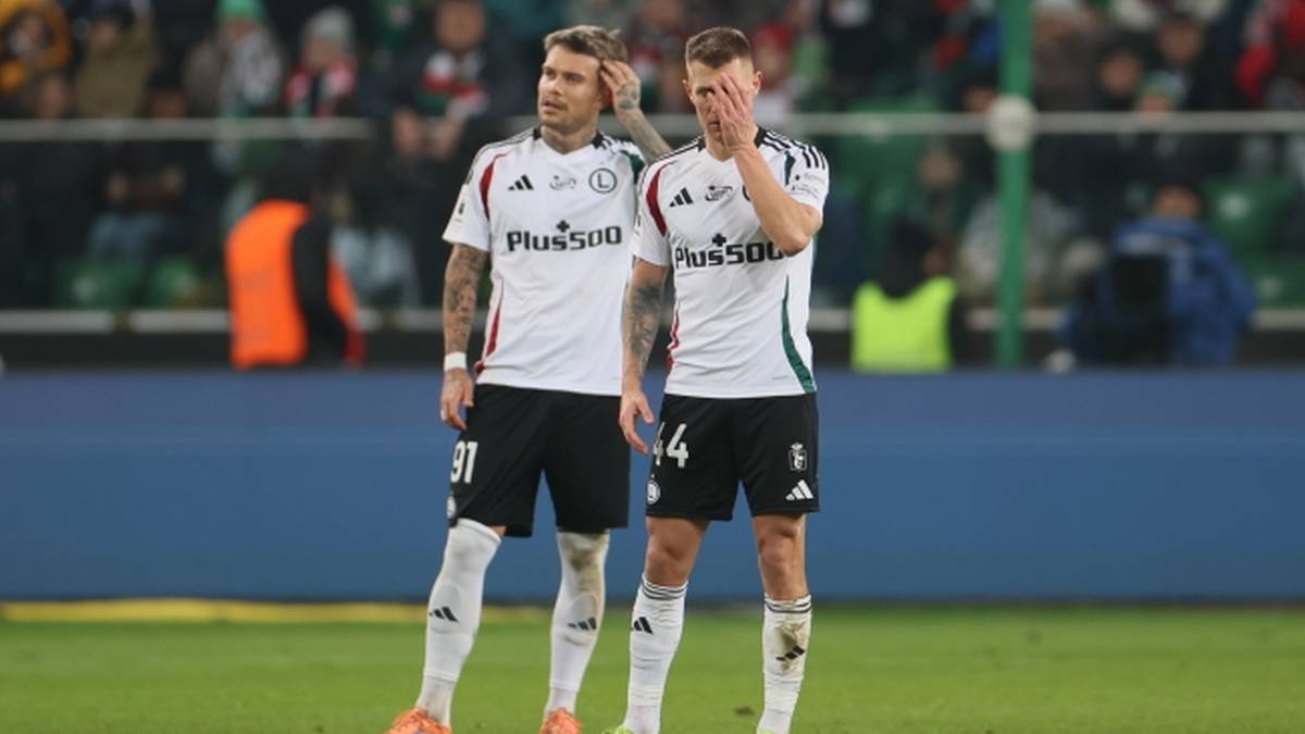 Dwóch piłkarzy Legii Warszawa w białych strojach meczowych, stojących na boisku, z wyrazem zmartwienia na twarzy.