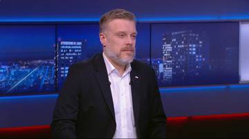 Adrian Zandberg w czarnym garniturze i białej koszuli, siedzi na tle ekranów z widokiem nocnego miasta.