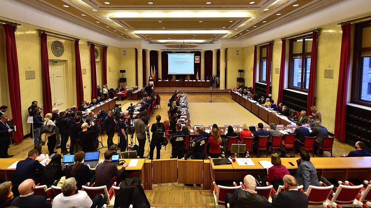 Sala obrad z licznymi uczestnikami siedzącymi przy stołach i na widowni, na środku duży ekran projekcyjny.