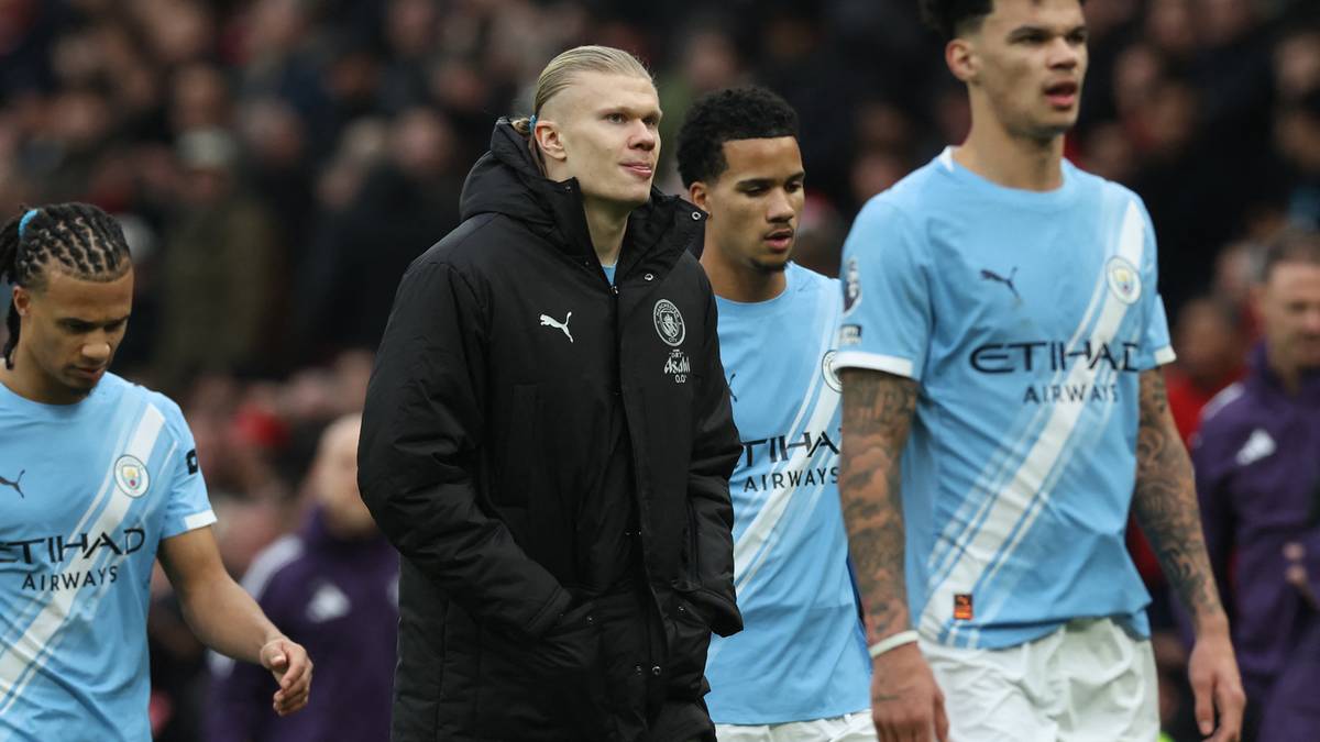 Piłkarze Manchesteru City w jasnych niebieskich strojach meczowych, jeden z nich w czarnej kurtce z kapturem, idą po boisku.