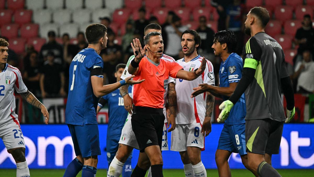 Sędzia w pomarańczowej koszulce stoi pośrodku grupy piłkarzy w niebieskich i białych strojach, gestykulując rękami. Pozostali zawodnicy zdają się dyskutować z nim lub między sobą.