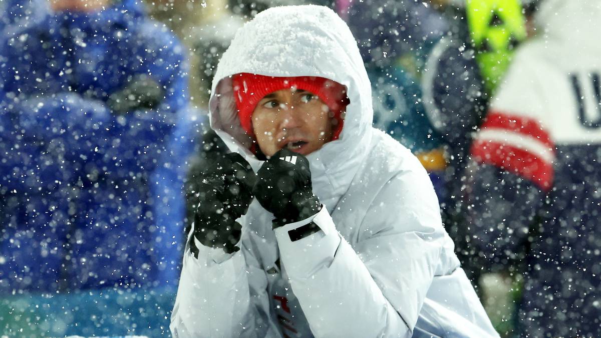 Skoczek narciarski w zimowym stroju, ubrany w białą kurtkę z kapturem i czerwoną czapkę, podczas zawod&oacute;w skok&oacute;w narciarskich.