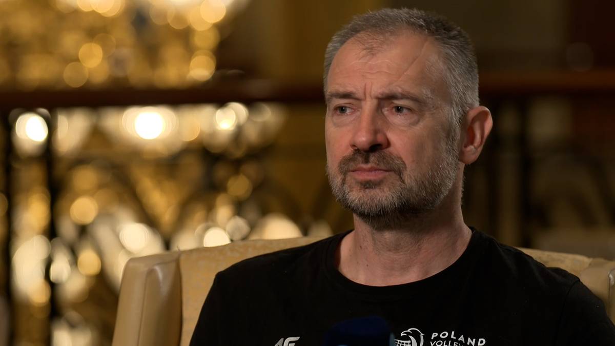 Portret mężczyzny z krótkimi, siwiejącymi włosami i kilkudniowym zarostem, ubranego w czarny t-shirt z napisem POLAND. Tło jest rozmyte, widoczne są złote i żółte światła.