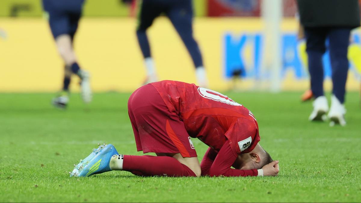 Piłkarz w czerwonej koszulce klęczy na boisku z głową ukrytą w dłoniach, w tle inni zawodnicy i trener.