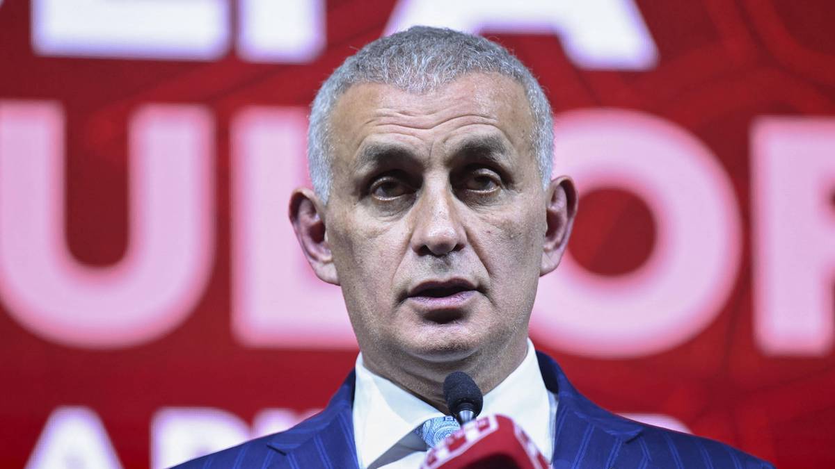 Portret mężczyzny w garniturze przemawiającego do mikrofonu, z czerwonym tłem z napisem.