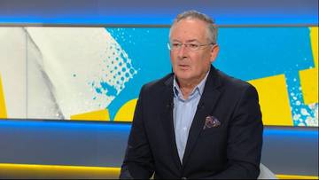 Bartłomiej Sienkiewicz w studio telewizyjnym, ubrany w ciemny garnitur i błękitną koszulę, z okularami na nosie, patrzy w stronę kamery.