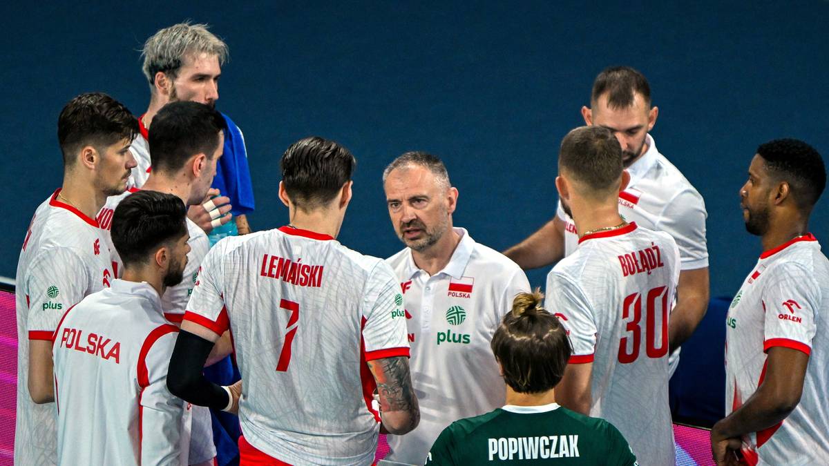 Grupa siatkarzy w białych strojach z czerwonymi numerami i napisami, otoczona wok&oacute;ł trenera. Wszyscy patrzą na trenera, słuchając go uważnie. Scena sugeruje moment analizy taktycznej lub odprawy drużyny.