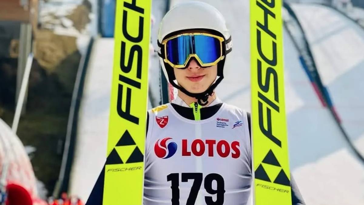 18-letni skoczek narciarski w białym kasku i goglach narciarskich, ubrany w biały kombinezon z numerem 179 i logiem LOTOS, stoi między dwoma żółtymi nartami z napisem FISCHER.