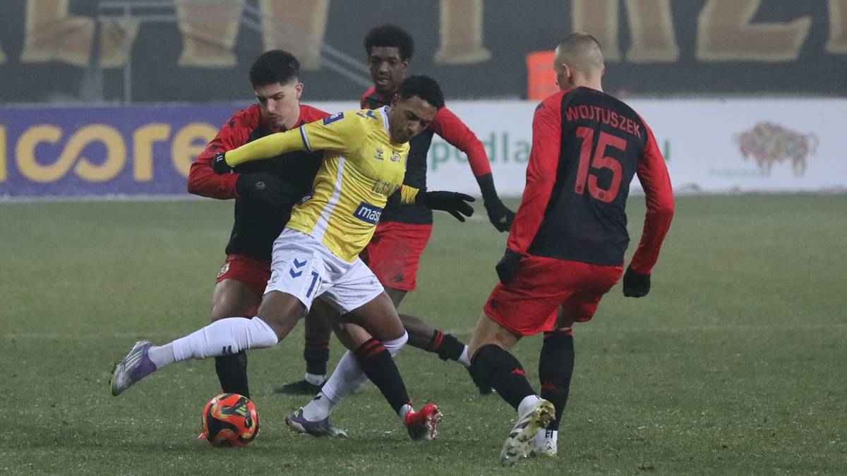 Piłkarz w ż&oacute;łto-białej koszulce walczy o piłkę z trzema przeciwnikami w czerwonych strojach.