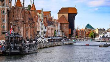 Historyczna kamienna zabudowa nad rzeką, z wielkim drewnianym żurawiem portowym w tle oraz zacumowanym żaglowcem. Ludzie spacerują po nabrzeżu.