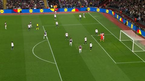 Widok z g&oacute;ry na boisko piłkarskie podczas meczu. Zawodnicy w białych i czarnych strojach rywalizują w okolicach bramki.