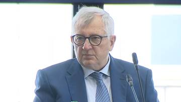 Starszy mężczyzna w garniturze siedzi przy stole z butelkami i mikrofonem.